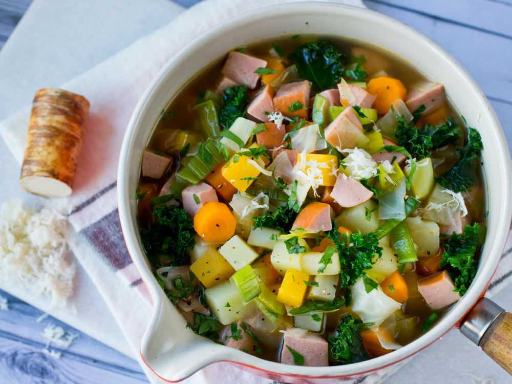 Hjemmelaget grønnsakssuppe med pølse i en hvit bolle, omgitt av et stykke brød og en skje på et hvitt bord.