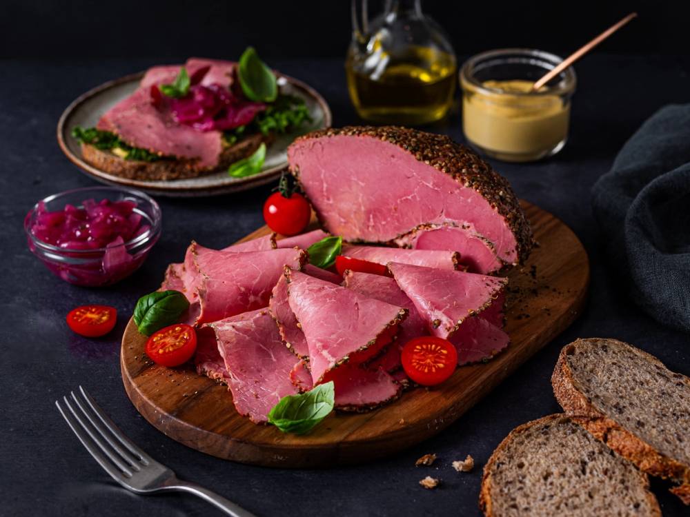 Skiver av kokt skinke på en rund treplate med cherrytomater og basilikum, omgitt av brødskiver, en liten skål med syltetøy, en liten bolle med rødkål, en glassflaske med olje og en liten skål med sennep på en svart bakgrunn.