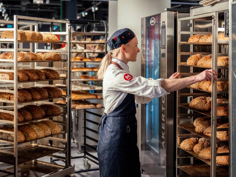 En baker i en bakeri som tar ut brød fra en ovn, omgitt av rader med ferdige brød på hyller.