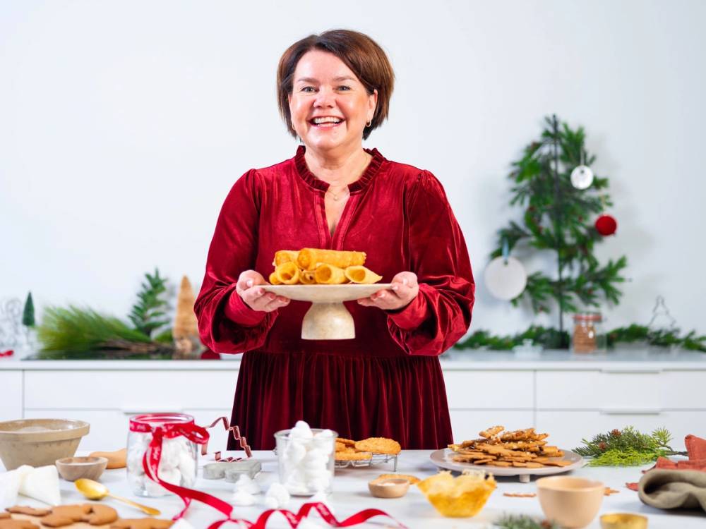 En kvinne i rød kjole holder et fat med kaker foran et juledekorert bord med juletrær i bakgrunnen.