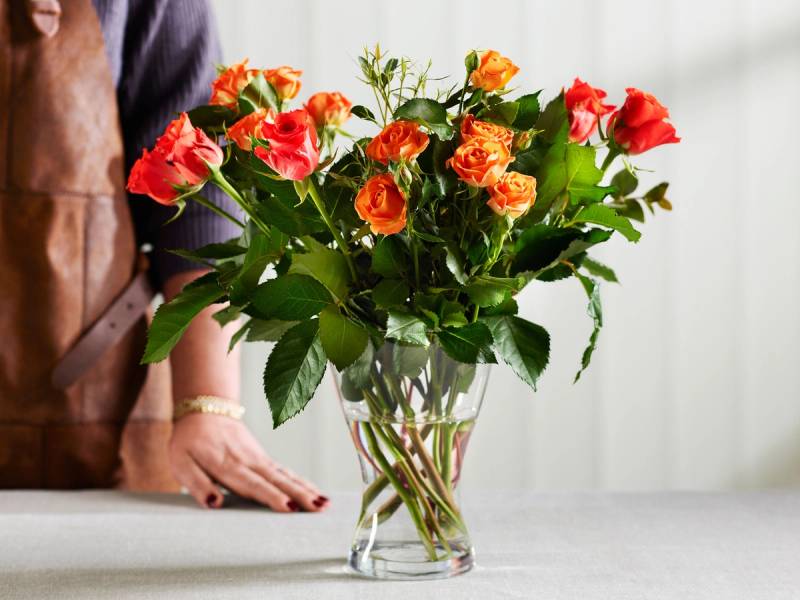 En person som står ved en bord med en stor vase fylt med snittblomster i røde og oransje nyanser.
