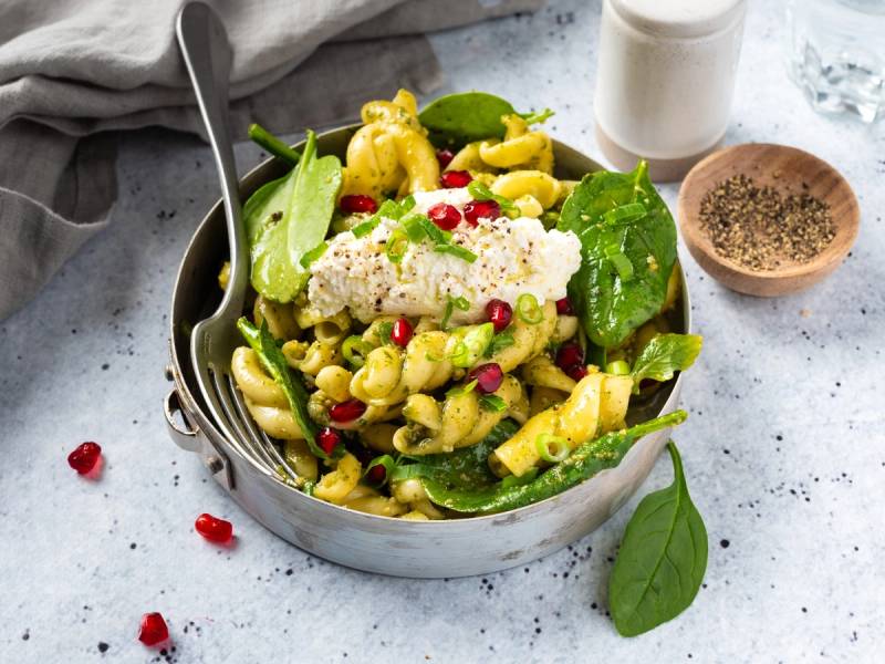 En pastasalat med pasta, spinat, fetaost og granateplekjerner i en bolle, med en skje i.