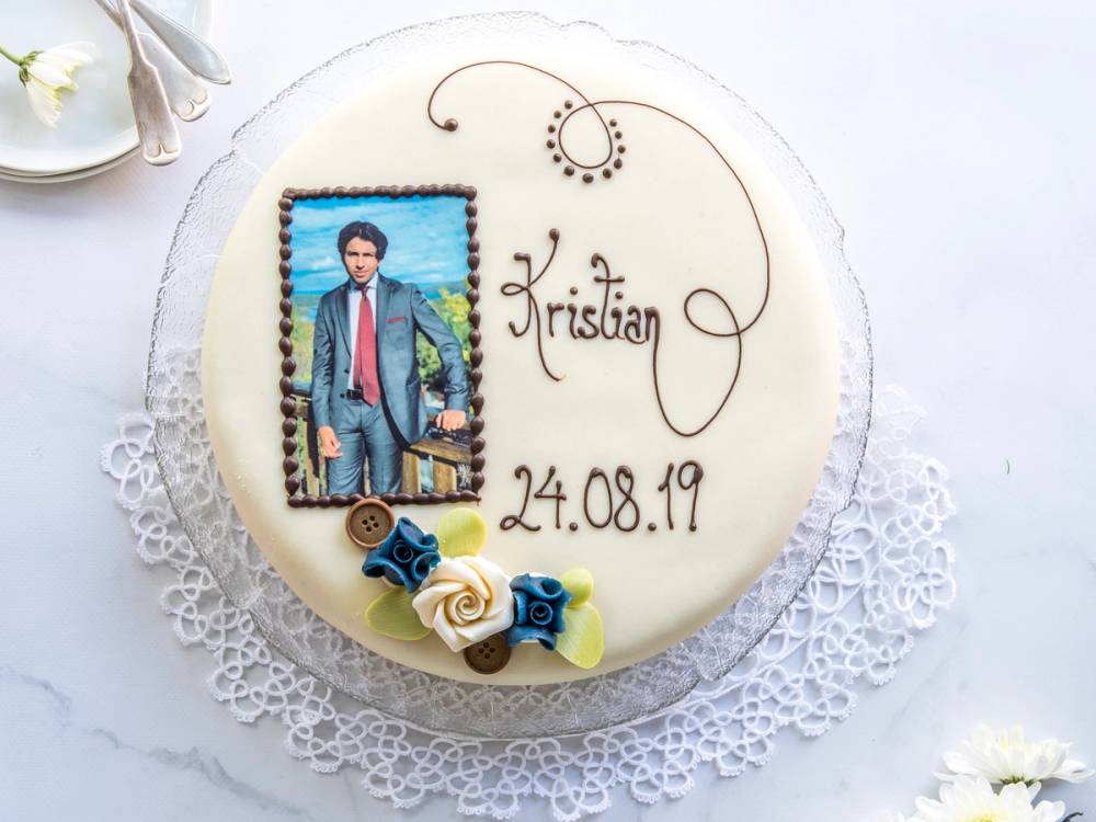En konfirmasjonskake med navnet Kristian og datoen 24.08.19, dekorert med en liten figur og blomster, på en hvit bordduk.