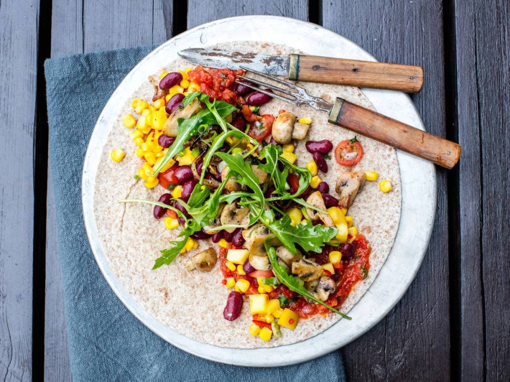 En tallerken med enchiladas, salsa, grønnsaker og salat, sett på et bord med en kniv og gaffel ved siden av.