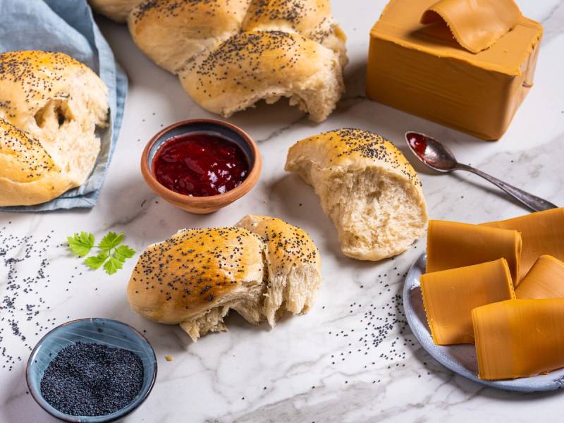 Fletteloff med poppyfrø, servert med en skål med jordbærsyltetøy og en kopp med gul ost, på et hvitt bord.