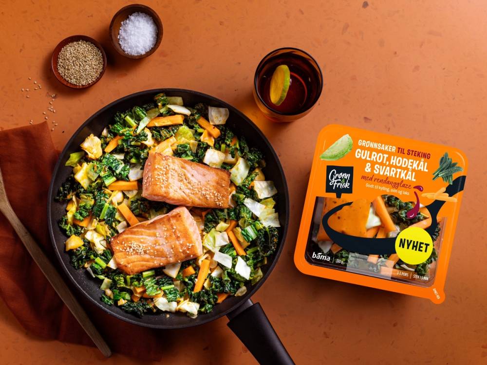 En stekepanne med laks og grønnsaker, en pakke med gulrot- og kålstaver, og krydder i små skåler på et bord.