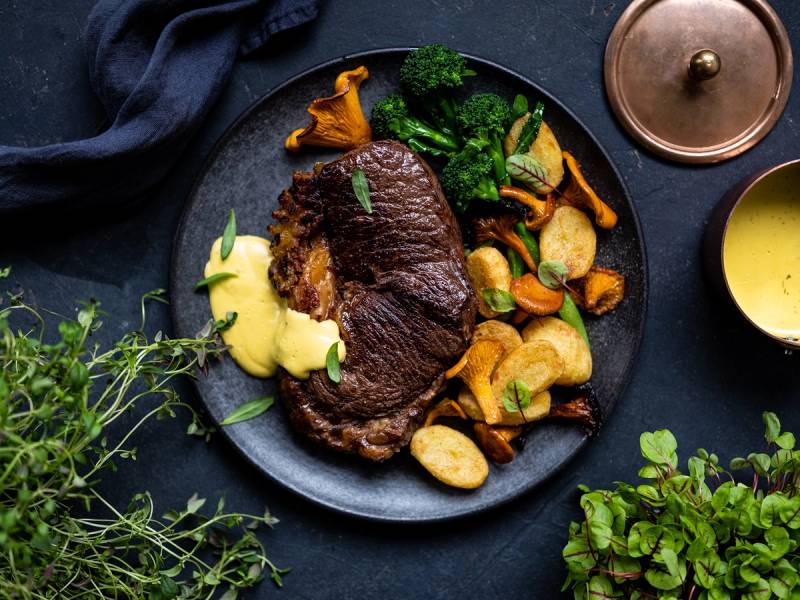 Stekt biff med ovnsbakte poteter og grønnsaker på en svart tallerken, med en liten saus eller sausflaske i bakgrunnen.