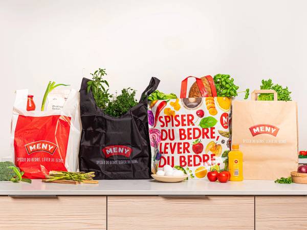 En rekke handleposer og matvarer på en kjøkkenbenk, inkludert en plastpose med frukt og grønnsaker, og en papirpose med grønne urter.