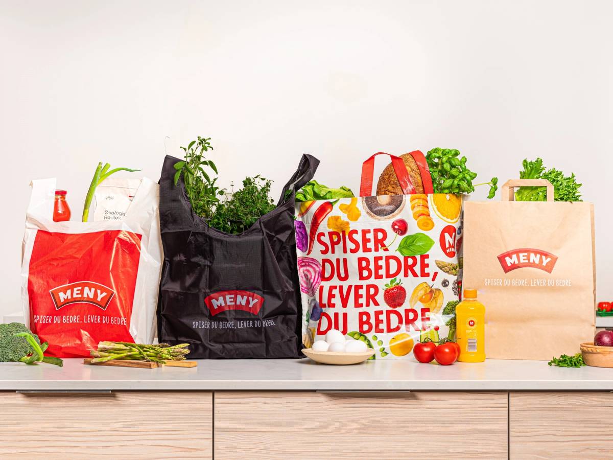 En rekke handleposer og matvarer på en kjøkkenbenk, inkludert en plastpose med frukt og grønnsaker, og en papirpose med grønne urter.