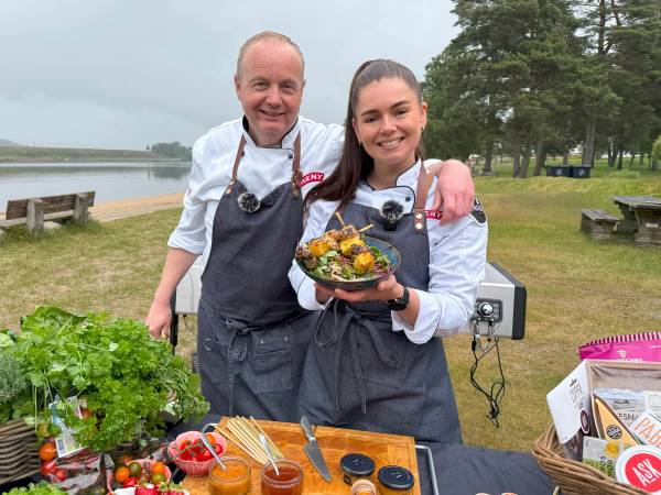 To personer i grillforkle står ved en utendørs grill med en skål salat, omgitt av grønnsaker og grillutstyr, med vann og trær i bakgrunnen.