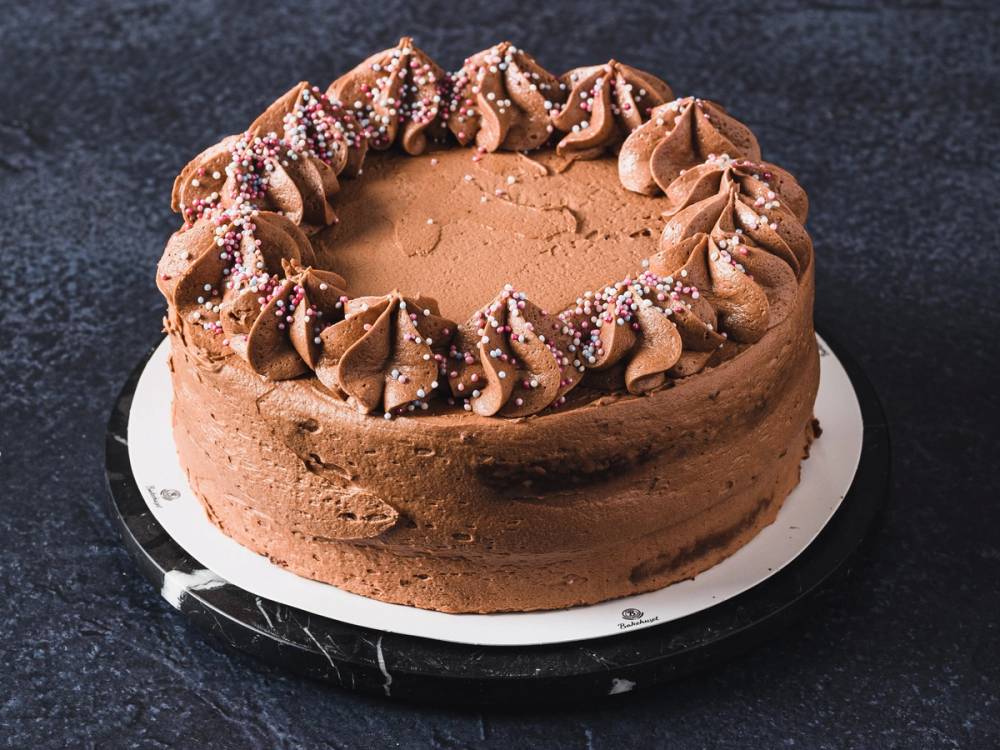 En rund sjokoladekake med krem og strøssel på toppen, plassert på en hvit kakeplate mot en mørk bakgrunn.