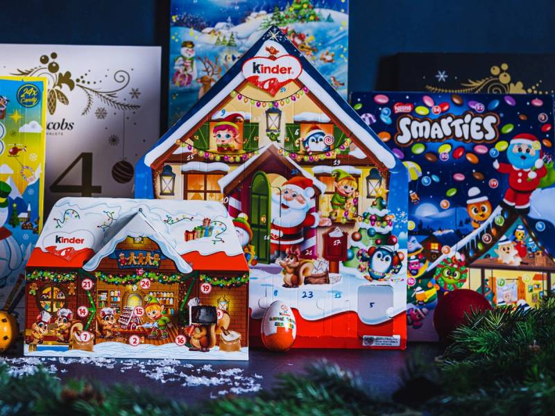 Ulike julekalendere med snø- og vintermotiv står oppstilt, inkludert en Kinder-kalender og en Smarties-kalender, med julepynt og granbar rundt.