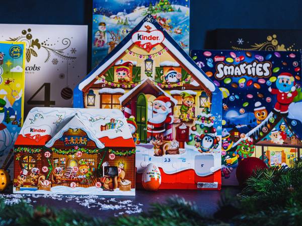 Ulike julekalendere med snø- og vintermotiv står oppstilt, inkludert en Kinder-kalender og en Smarties-kalender, med julepynt og granbar rundt.