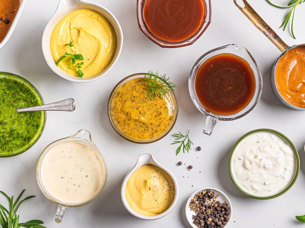 Ulike sauser og dressinger i små skåler på et hvitt bord, med noen grønne urter som pynt.
