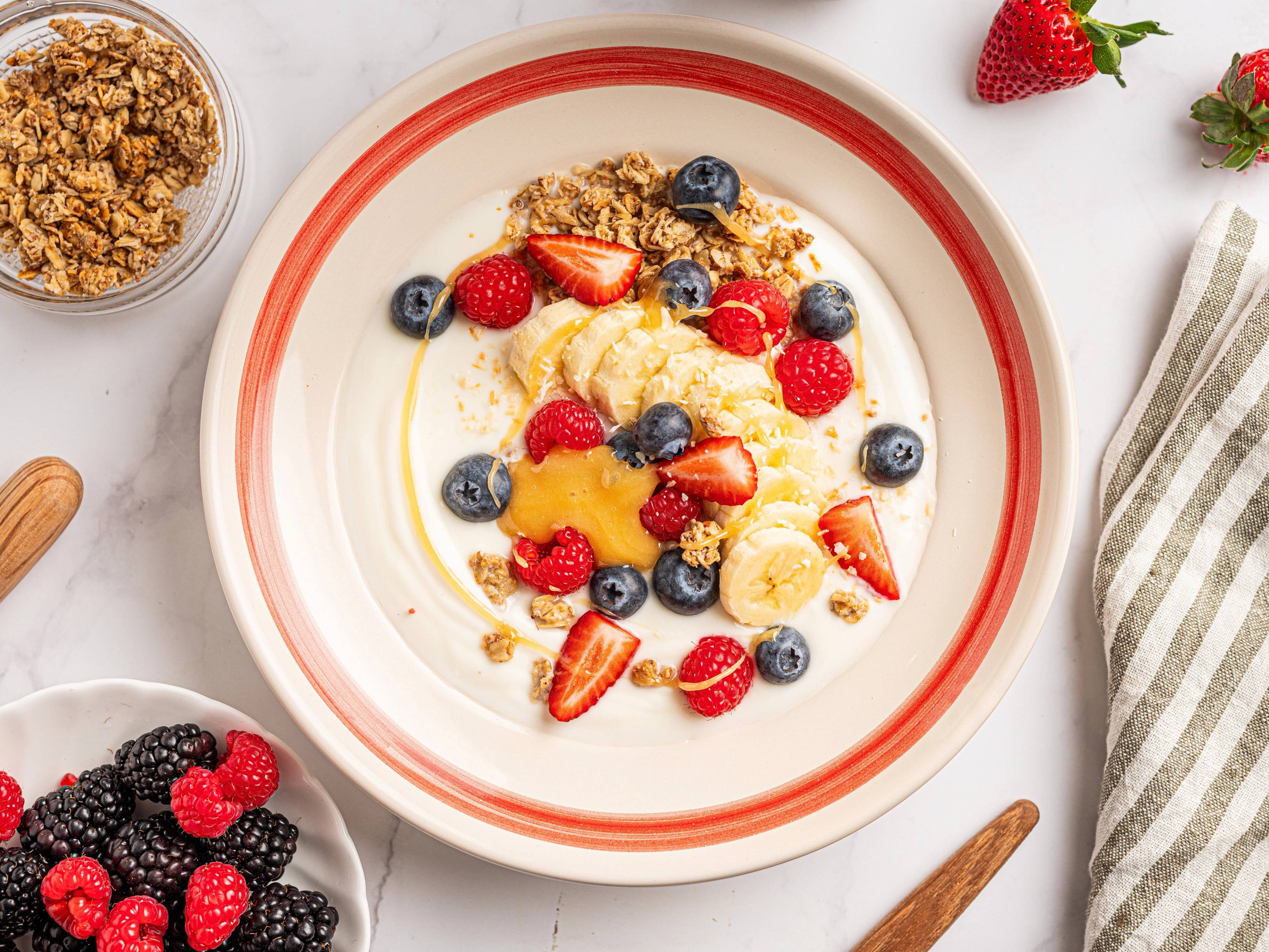 En yoghurtbolle med ferske jordbær, blåbær og granola på en hvit tallerken med rød kant.