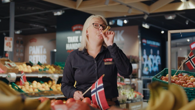 En person med blondt hår i en matbutikk holder en telefon til øret, omgitt av frukt og grønnsaker.