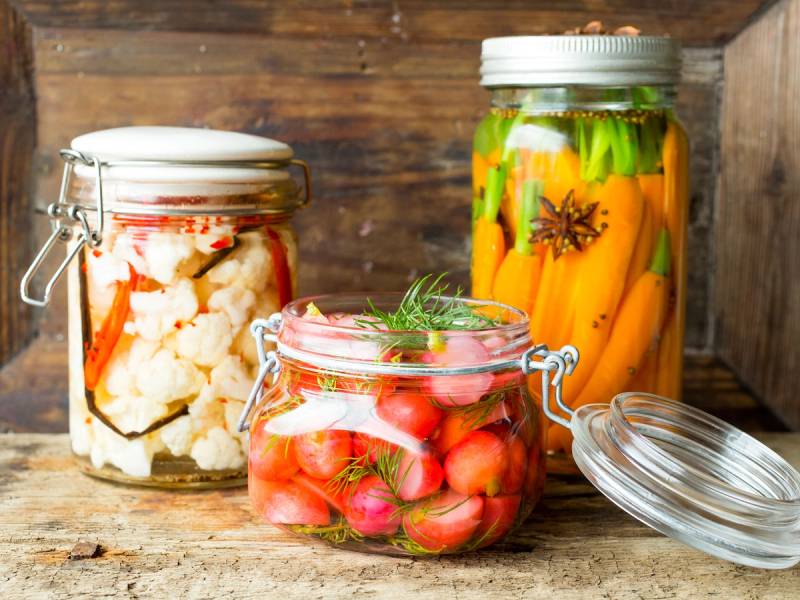 Syltede grønnsaker i glasskrukker på et treverk, inkludert blomkål, cherrytomater og gulrøtter med krydder.