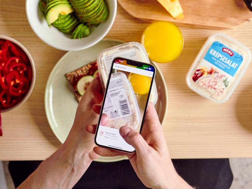 En person tar et bilde av en matrett med en smarttelefon, omgitt av ulike matvarer og drikke på et bord.