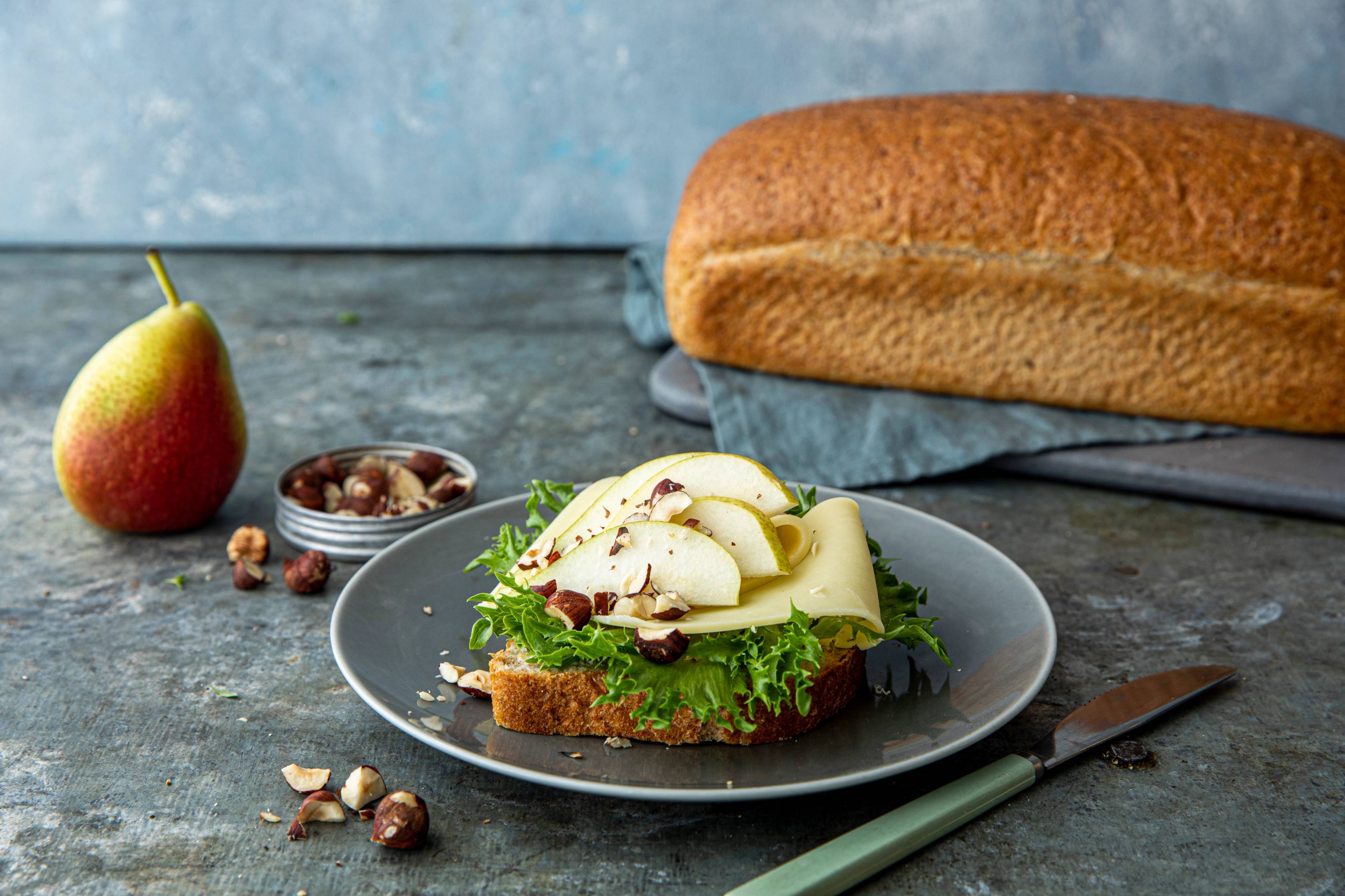 En åpen matpakke med grovt brød, ost, salat og noen nøtter, med et eple ved siden av, på en mørk tallerken og et kniv ved siden av.