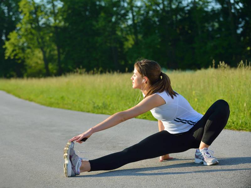 En person utfører tøying på en grusvei i et grønt landskap, med bena strukket ut til sidene og armene utstrakt foran seg.