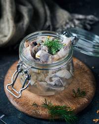 Sursild i glasskrukke med dill og sennepsfrø på et rundt treplate, klar til servering.