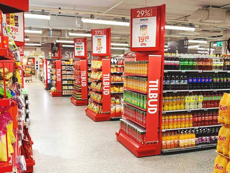 Aisle in a grocery store with shelves filled with various beverages and promotional signs hanging from the ceiling.
