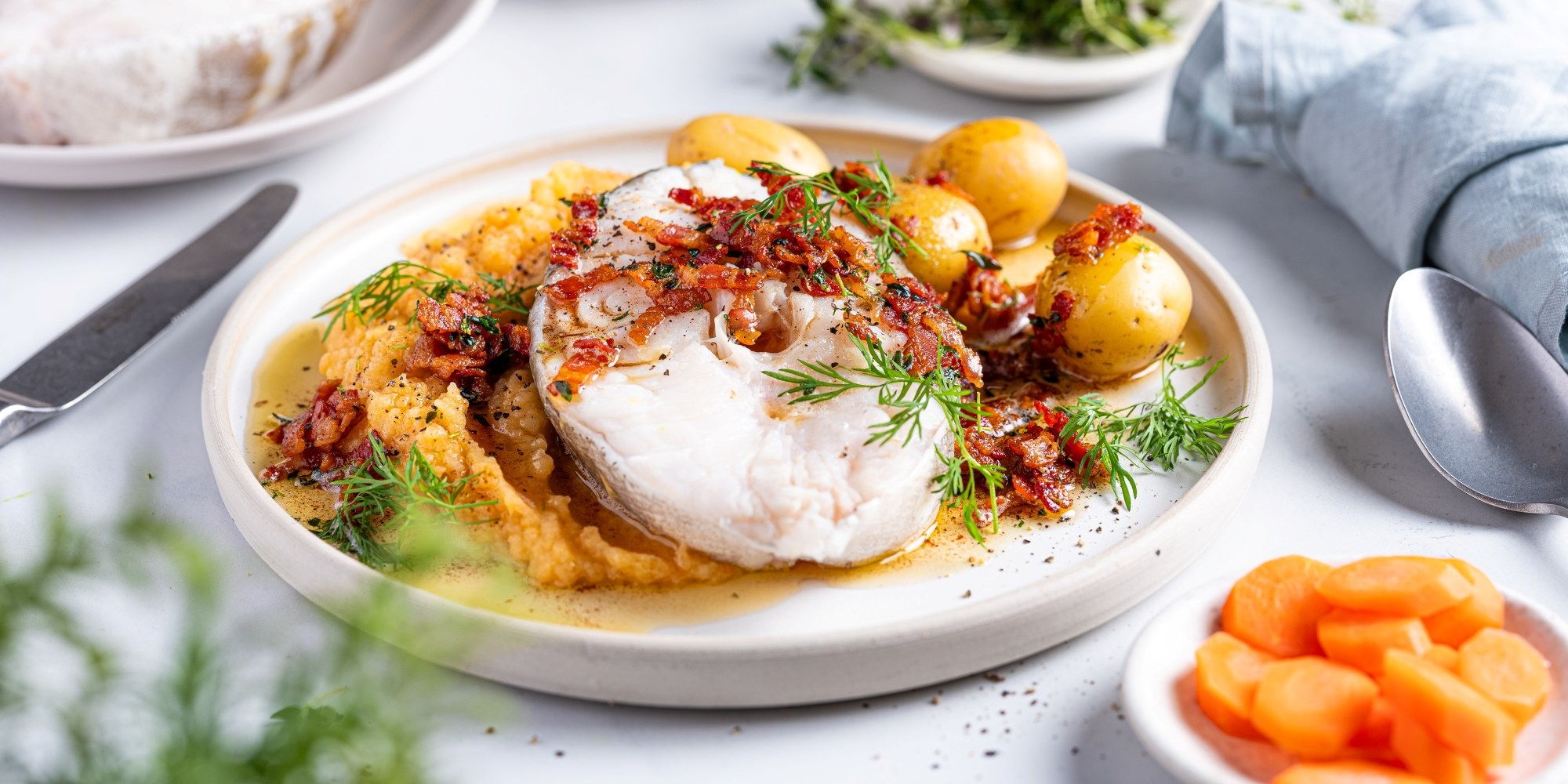 Skrei med rotmos og små poteter på en hvit tallerken, servert med grønne urter og en skje ved siden av.