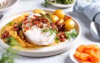 Skrei med rotmos og små poteter på en hvit tallerken, servert med grønne urter og en skje ved siden av.