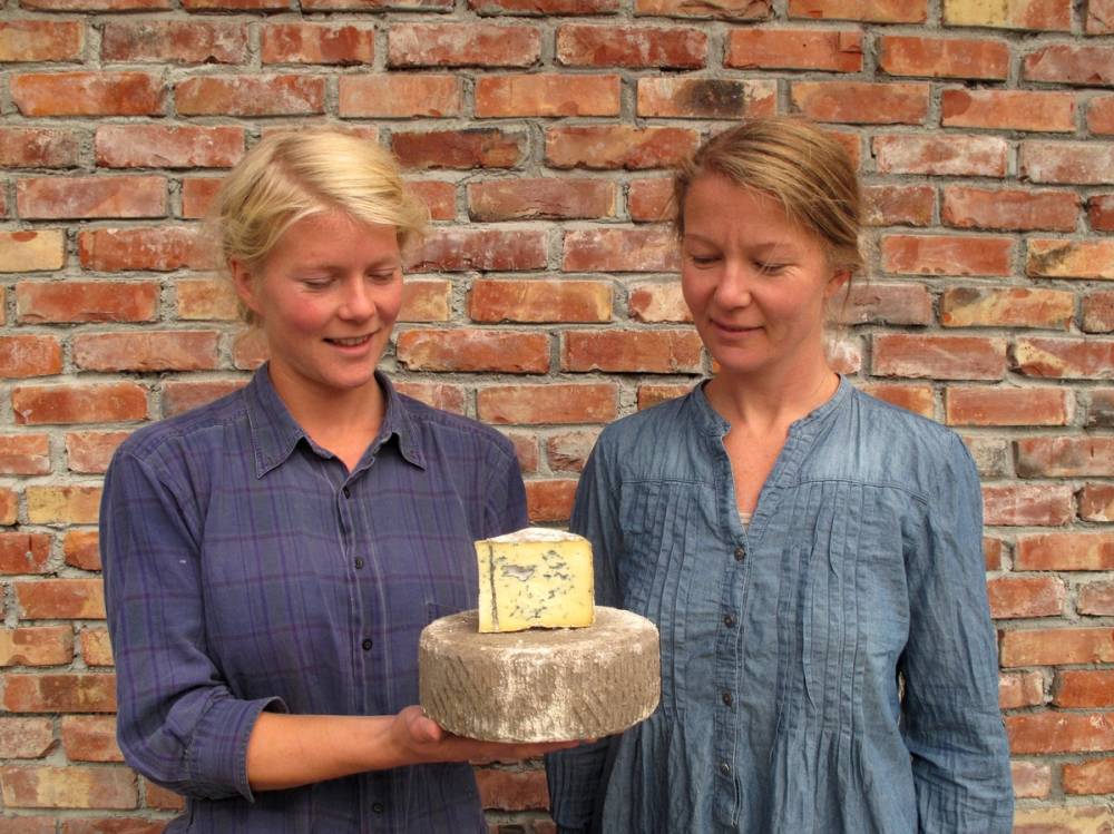 To kvinner står side om side foran en murvegg, og holder et kakefat med en kake på, uten å vise ansiktene tydelig.