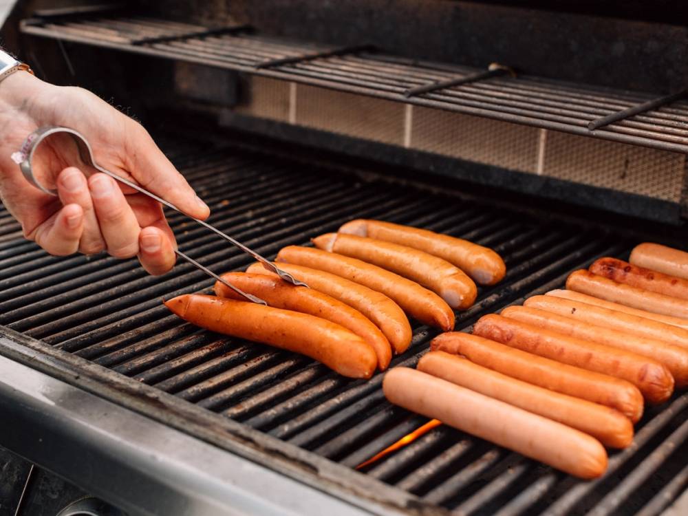 En person bruker pinsett for å legge pølser på grillen, hvor flere pølser allerede er grillet.