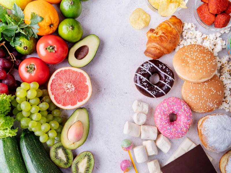 Et utvalg av frukt, grønnsaker og søtsaker på et bord, inkludert tomater, avokado, druer, grapefrukt, croissant, donuts og marshmallows.