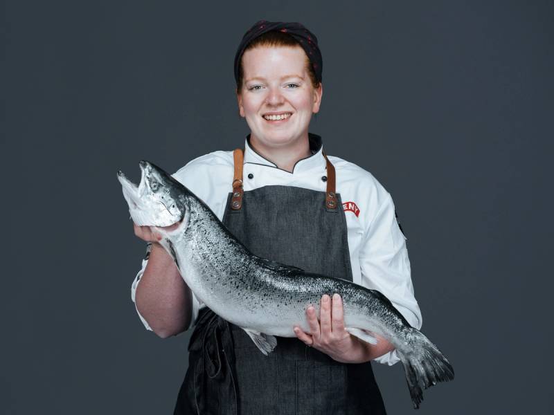 En person i kokkeforkledning holder en stor ørret foran en mørk bakgrunn.