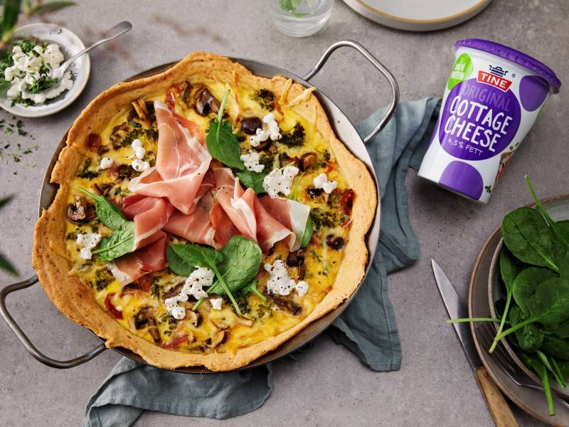 En pizza med cottage cheese, skinke og grønne blader på en tallerken, sett på et bord med bestikk og en kopp, klar til å spises.