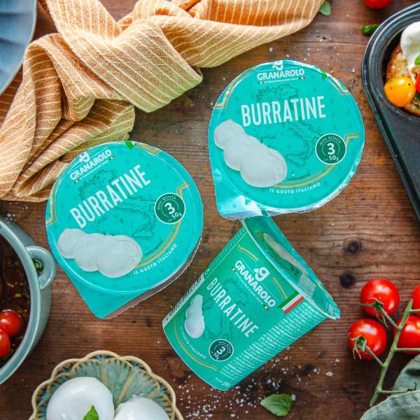 To pakker med Granarolo burrata ost og en kopp med en grønn stripe, omgitt av tomater og en skål med salat, på et trebord.