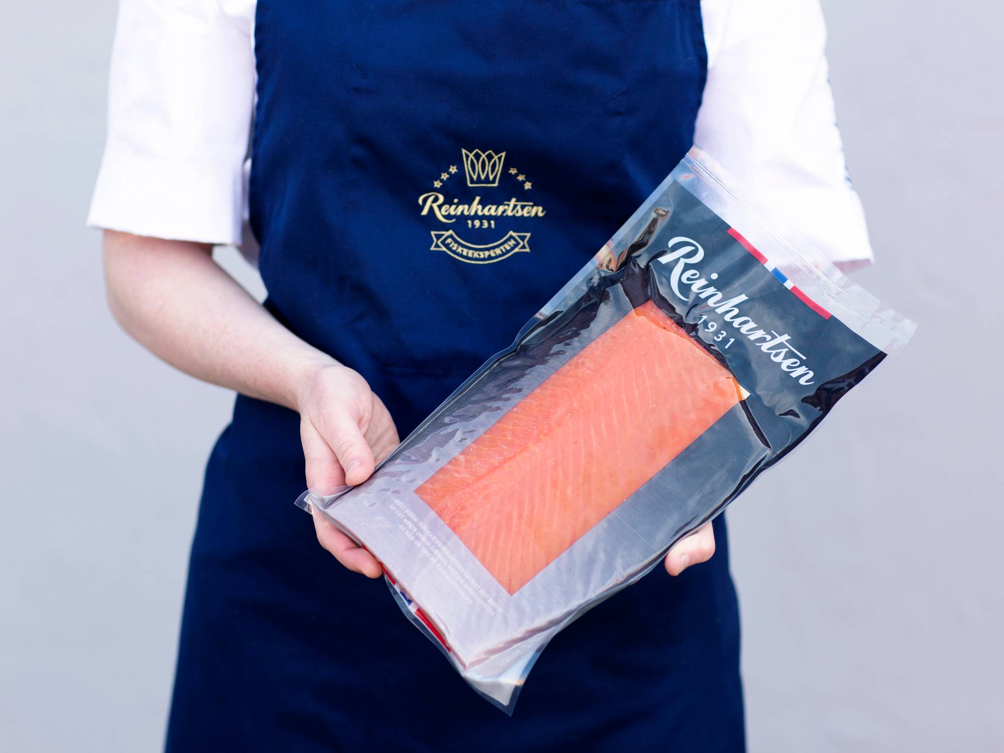 Person i blå apron som holder en pakke med laks, med Reinhartsen-logo på forpakningen.