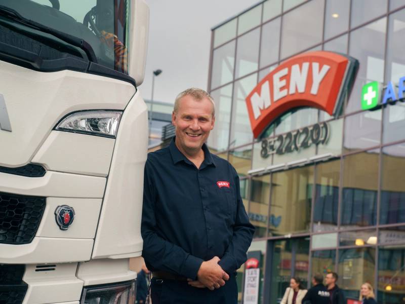 En person står foran en stor lastebil uten å vise ansikt, med et kjøpesenter i bakgrunnen.