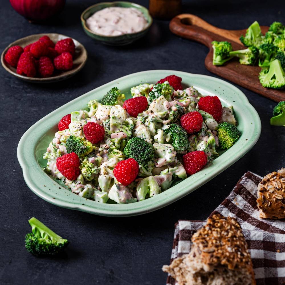 En salat med brokkoli, reddiker og dressing på en oval grønn tallerken, omgitt av frukt, grønnsaker og brød på en mørk bakgrunn.