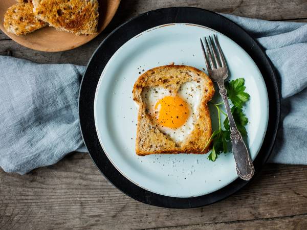 Et stykke brød med et egg stekt i midten, servert på en hvit tallerken med en liten grønn urt og en gaffel ved siden av.