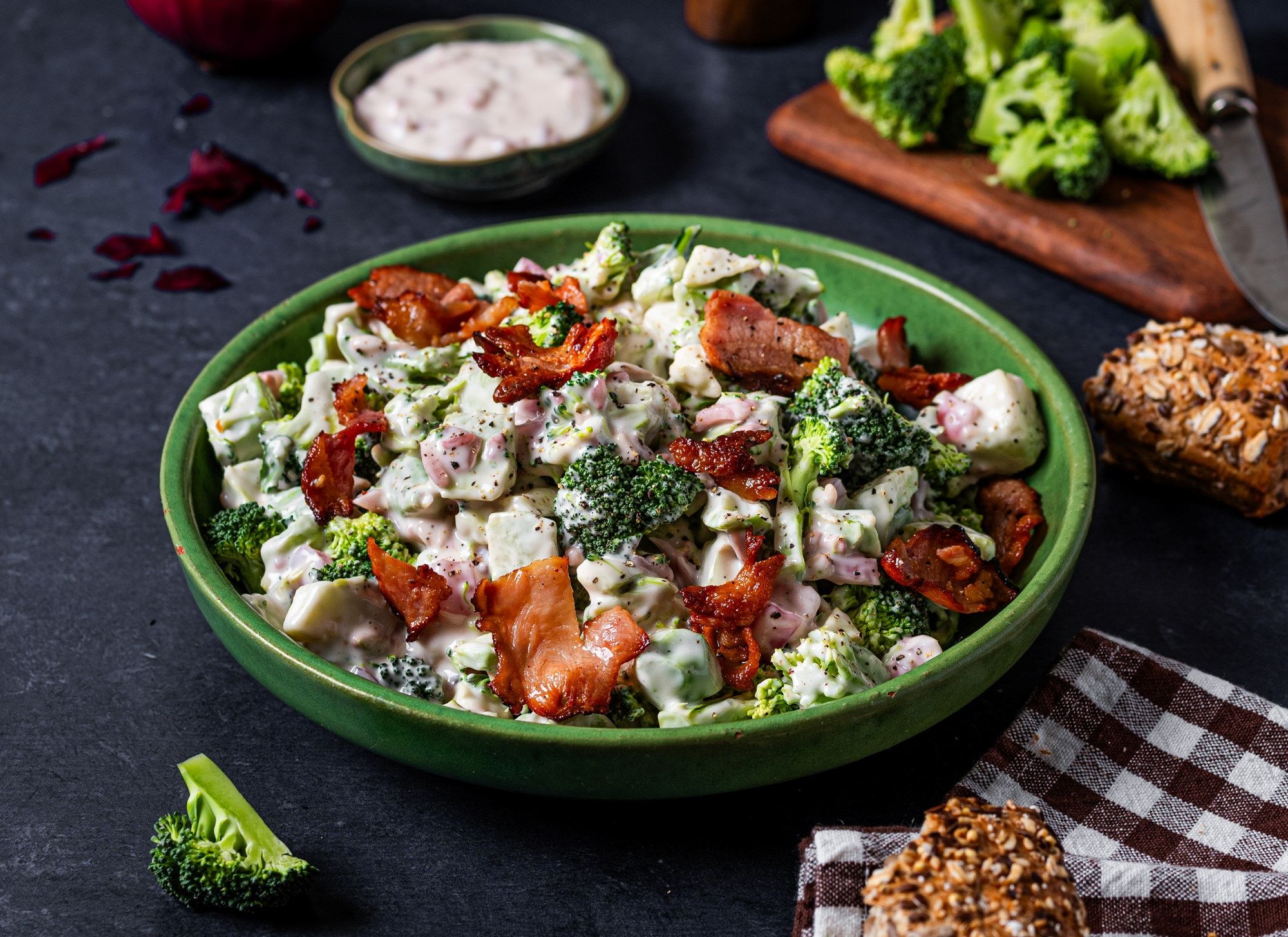 En stor grønn bolle med en salat som inneholder brokkoli, tomater, ost, bacon og nøtter, plassert på et mørkt bord med en liten skål med dressing og en skjærefjøl med grønnsaker i bakgrunnen.