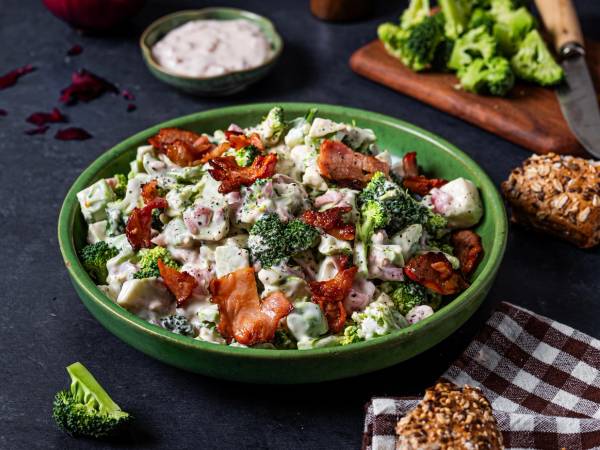En stor grønn bolle med en salat som inneholder brokkoli, tomater, ost, bacon og nøtter, plassert på et mørkt bord med en liten skål med dressing og en skjærefjøl med grønnsaker i bakgrunnen.