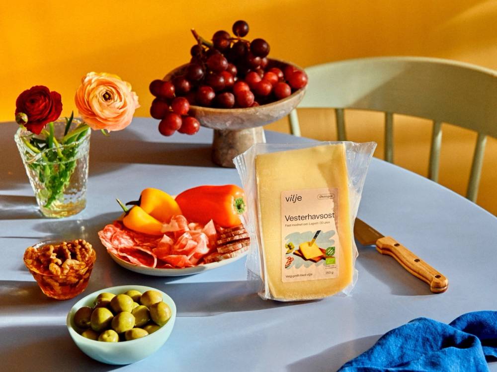 Et bord dekket med en skål med druer, en pakke med ost, en tallerken med spekemat og frukt, en liten bolle med oliven, og en vase med blomster, med en gul bakgrunn og en grønn stol.