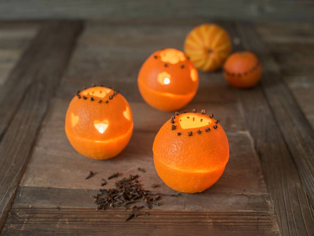 Tre klementiner med lysende lys inni, plassert på et trebord med noen krydder eller frø rundt.
