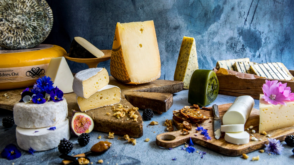 Et utvalg av ulike oster, noen med tilbehør som frukt og kjeks, på et bord med en blå bakgrunn.