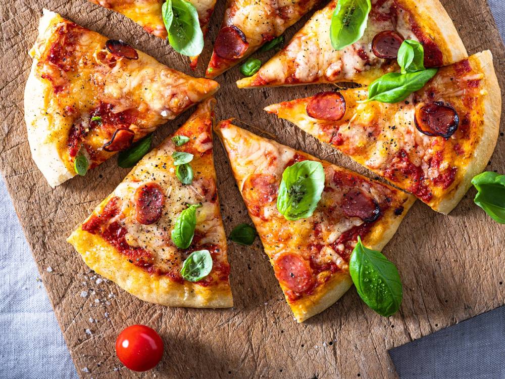 Pizza med pepperoni, grønne basilikumblader og smeltet ost på en treplate, med en enkelt cherrytomat ved siden av.