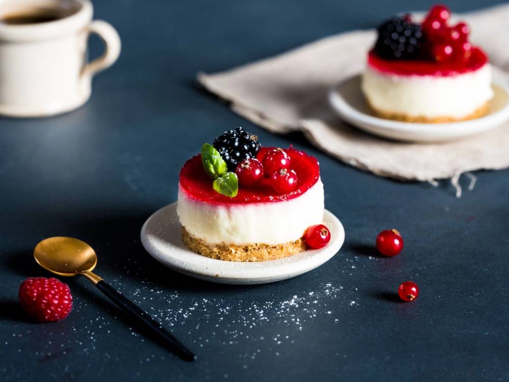 To små ostekaker med bær og mynte på en hvit tallerken, omgitt av noen friske bær og en skje, på en mørk bordflate.