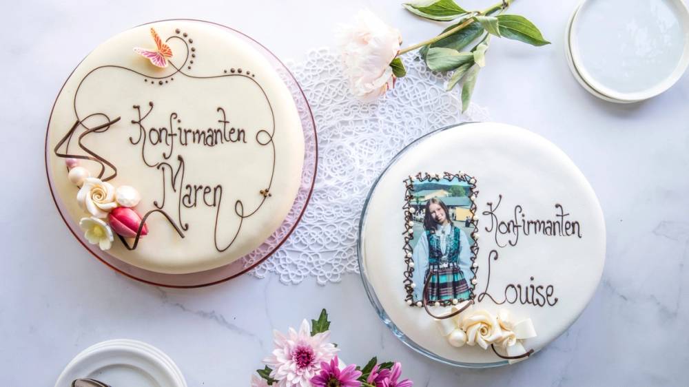 To konfirmasjonskaker med dekorasjoner og navn, omgitt av blomster og servise på et bord.