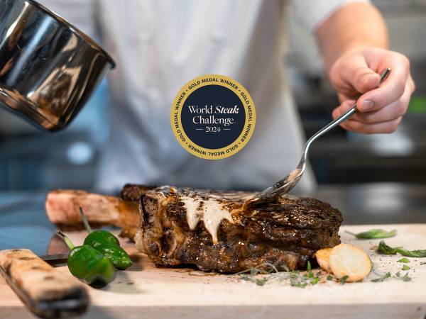 En person skjærer i en stor, grillet biff på et kjøkkenbord med grønnsaker og urter rundt.