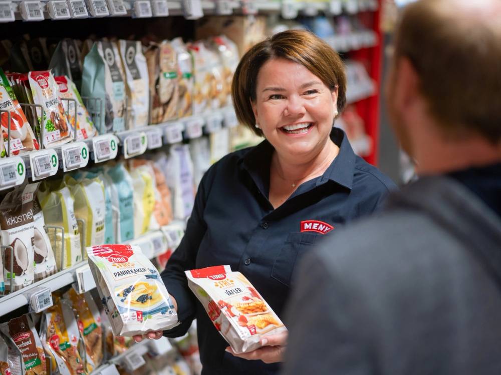 En butikkansatt hjelper en kunde med å velge melk i en dagligvarebutikk, med hyller fylt av ulike melkekartonger i bakgrunnen.