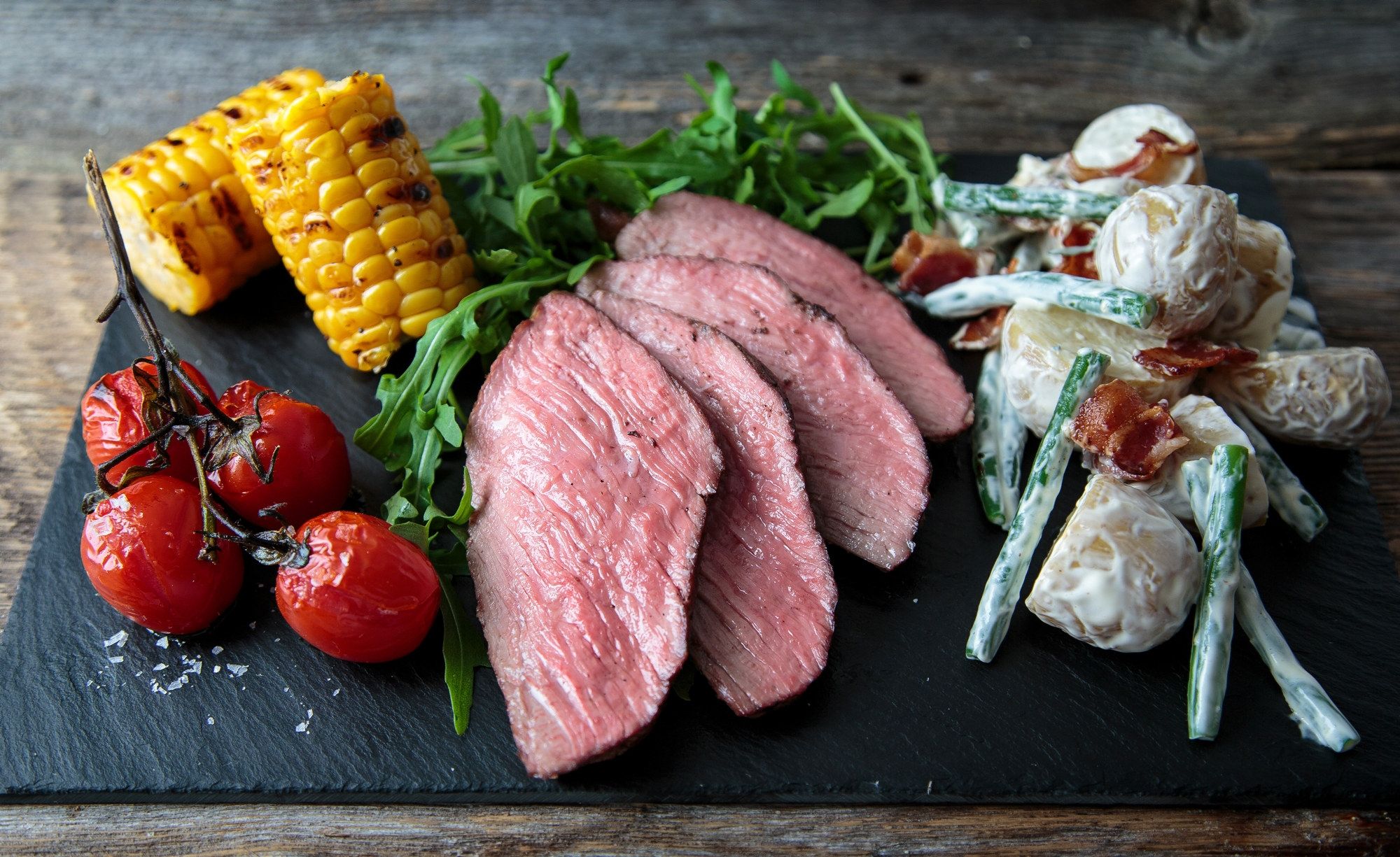 Servering av stekt biff med grillede maiskolber, cherrytomater, salat og potetsalat på en svart skjærefjøl.