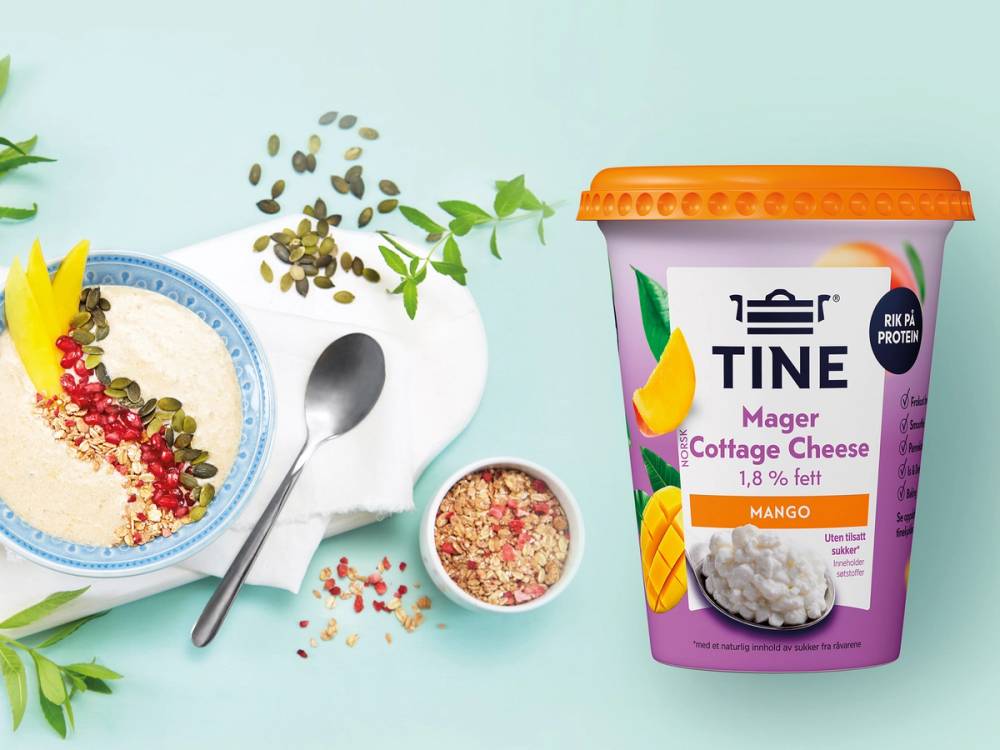 Smoothie bowl med cottage cheese og mango, omgitt av frø og urter, med en kopp og skje ved siden av.